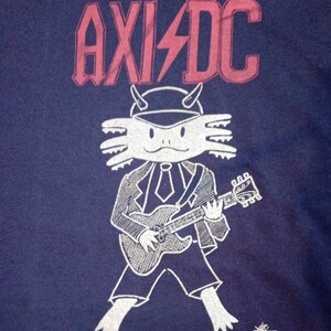 May include: A blue t-shirt featuring a white axolotl wearing a suit and playing a guitar. The text "AXI/DC" is printed in red above the axolotl.