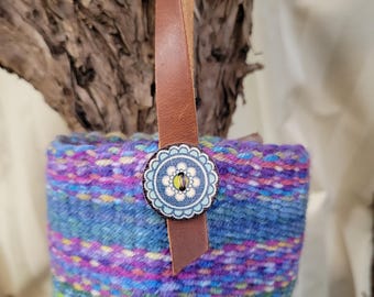 Può includere: Un portabicchieri colorato e fatto a mano con un cinturino in pelle e un bottone decorativo. Il portabicchieri è realizzato in tessuto intrecciato nei toni del blu, viola, verde e giallo. Il bottone ha un motivo floreale azzurro con un centro giallo.