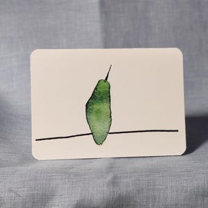 May include: A white rectangular card with rounded corners features a watercolor illustration of a green pear shape perched on a thin black line. The pear has a dark green stem. The card is set against a light blue fabric background.