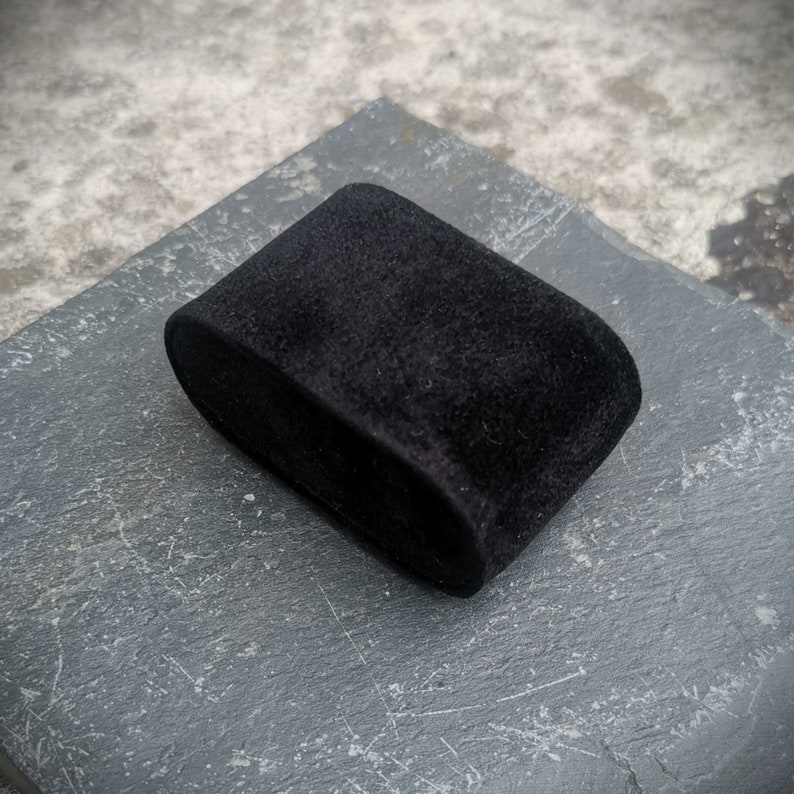 K&ouml;nnte beinhalten: Eine schwarze Samt-Ringbox mit abgerundeter Form. Die Box steht auf einer grauen, strukturierten Oberfl&auml;che.
