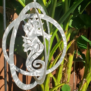 May include: A silver metal wind chime featuring a seahorse design. The seahorse is surrounded by three concentric circles. The wind chime is hanging from a string in front of a green leafy plant.