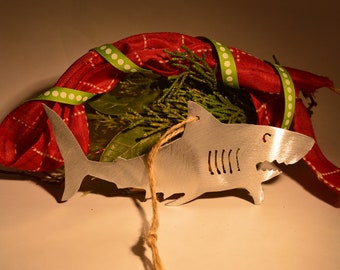 May include: A silver shark ornament with a string for hanging. The ornament is surrounded by red and green fabric with white polka dots.