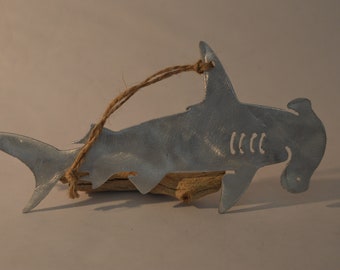 May include: A silver metal hammerhead shark ornament with a brown twine hanger. The ornament is resting on a piece of driftwood.