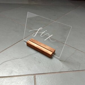 May include: A clear acrylic sign with the word "six" written in white calligraphy on a wooden stand.