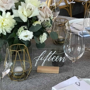 May include: A white acrylic table number with the word "fifteen" written in black calligraphy. The table number is on a wooden base and is sitting on a gray tablecloth. There are two gold geometric candle holders and a glass of water on the table.