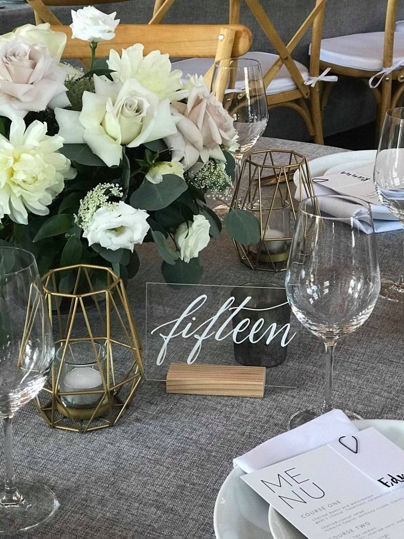 May include: A white acrylic table number with the word "fifteen" written in black calligraphy. The table number is on a wooden base and is sitting on a gray tablecloth. There are two gold geometric candle holders and a glass of water on the table.