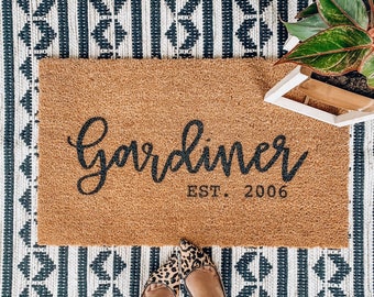 May include: A rectangular coir doormat with the name "Gardiner" in black script and "EST. 2006" printed below. The doormat is on a patterned rug with a black and white geometric design. A potted plant is visible in the upper right corner.