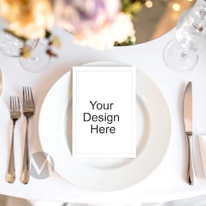 May include: A white place setting with a blank card that reads "Your Design Here" on a white plate. Silverware, including a fork and knife, is arranged on either side of the plate. The background includes flowers and glassware.