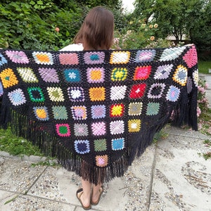 Può includere: Uno scialle nero all'uncinetto con motivi quadrati colorati e frange. Lo scialle è fatto di filato e ha una varietà di colori, tra cui rosso, verde, blu, giallo e rosa.