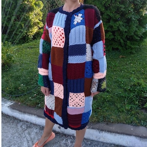 May include: A long-sleeved, knee-length cardigan constructed from a patchwork of various knitted and crocheted squares in diverse colours including navy blue, burgundy, light blue, pink, brown, and green, featuring a round neckline and front button closure.