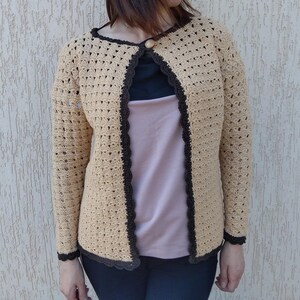 Puede incluir: Un cárdigan de ganchillo beige con un patrón calado repetitivo, mangas largas y un borde festoneado de color marrón oscuro. La prenda se cierra en el escote con un único botón redondo y se lleva sobre una camiseta superpuesta.