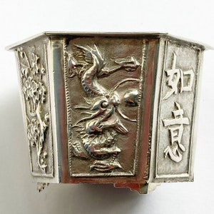 May include: A silver hexagonal box with a dragon design and Chinese characters. The box has a raised rim and a decorative base.
