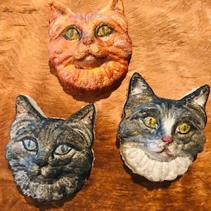 Könnte beinhalten: Drei Keramik-Katzenkopfskulpturen. Die erste Katze ist orange mit einem weißen Fleck auf der Brust. Die zweite Katze ist grau mit weißem Fell um den Hals. Die dritte Katze ist schwarz-weiß mit einem weißen Fleck auf der Brust.