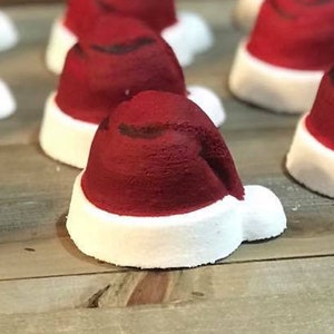 May include: A collection of red and white bath bombs shaped like Santa hats. The bath bombs are arranged in a row on a wooden surface.