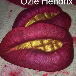 May include: Three bath bombs shaped like lips with a gold glitter design. The bath bombs are red and have a gold glitter design.