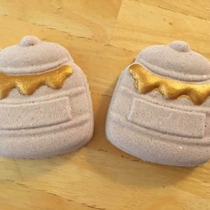 Puede incluir: Dos bombas de baño moradas con forma de tarro de miel con miel dorada goteando en la parte superior.