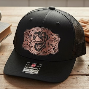 Puede incluir: Gorra de camionero negra con un diseño de perro color cobre en la parte delantera. La gorra tiene una visera curva y una parte trasera de malla. Una taza de café y libros están en el fondo.