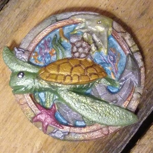 May include: A circular, green, gold, and silver resin sculpture of a sea turtle swimming in a circle with a starfish, seashells, and coral. The sculpture is set against a wooden background.