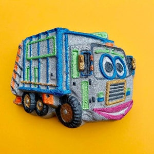 Może przedstawiać: Zabawkowy szary i niebieski samochód śmieciarski z uśmiechającą się twarzą, wypukłymi oczami i różowymi ustami. Samochód ma zielono-białe paski z boku i złoty grill.