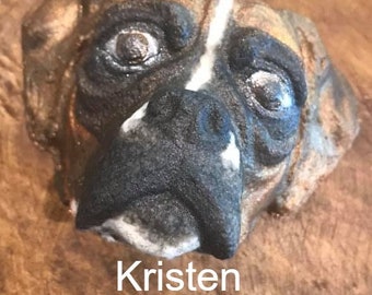 May include: Close-up of a ceramic dog sculpture with brown, black, and white paint. The sculpture is of a boxer dog's head with a white stripe on its nose. The sculpture is signed "Kristen Benjamin".
