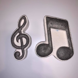 Puede incluir: Dos bombas de baño blancas con forma de clave de sol y una nota musical. Las bombas de baño tienen contornos negros.