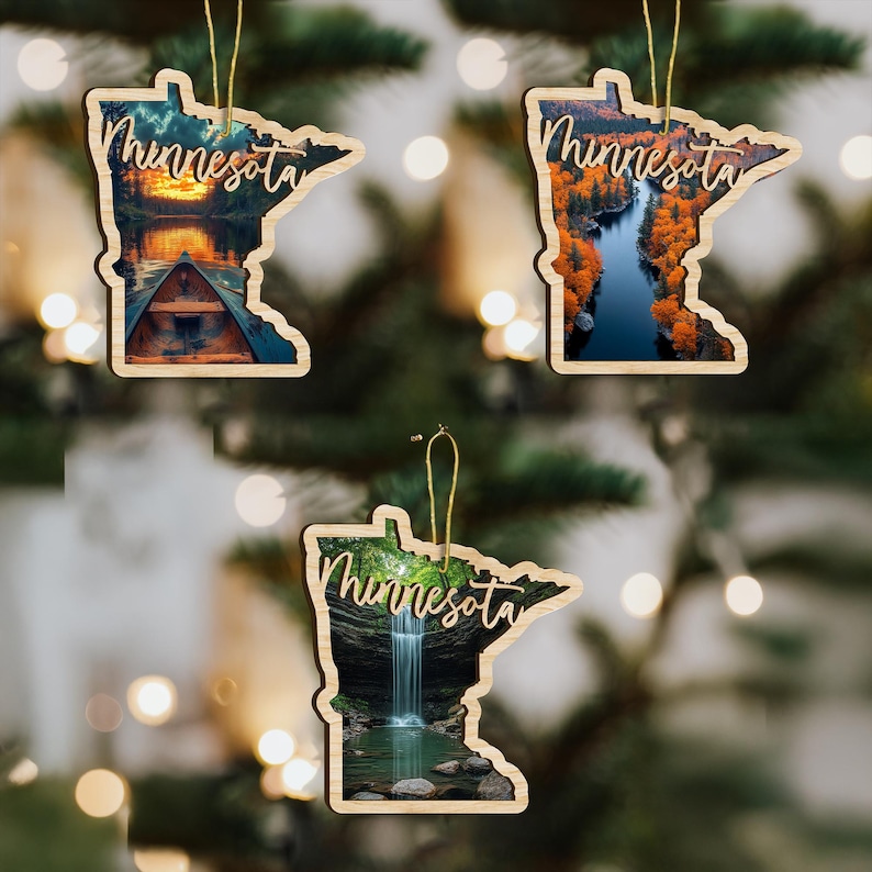 May include: Three wooden Minnesota state-shaped ornaments with scenic landscape images. Each ornament has the word "Minnesota" in script. One shows a cabin, another a river with fall foliage, and the third a waterfall.