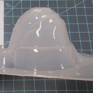 Op de afbeelding: Een transparante plastic mal in de vorm van een heksenhoed. De mal is ontworpen om een hoed te maken met een brede rand en een hoge, puntige kroon.