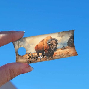 May include: A brown and white bison with a name tag that says "Joseph" on it. The tag is a light brown colour with a watercolour background of a grassy field.