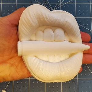 Pu&ograve; includere: Scultura in ceramica bianca di una bocca che tiene un proiettile tra i denti. La scultura &egrave; tenuta nella mano di una persona.
