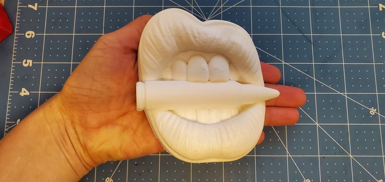 Pu&ograve; includere: Scultura in ceramica bianca di una bocca che tiene un proiettile tra i denti. La scultura &egrave; tenuta nella mano di una persona.