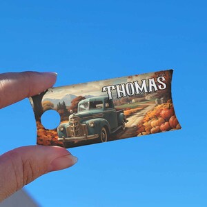 May include: A blue vintage pickup truck drives down a country road lined with autumn foliage and pumpkins. The truck has the name "THOMAS" printed on the side.