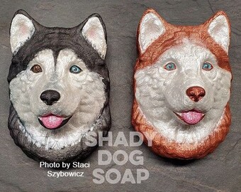 May include: Two handmade soap bars shaped like husky dog faces. The soap on the left is black and white, and the soap on the right is copper and white. Both soaps have a shiny finish and are made by Shady Dog Soap.
