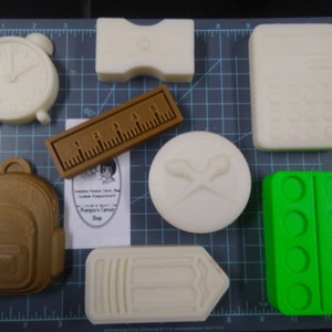 Puede incluir: Un conjunto de nueve moldes de silicona para hacer varios artículos con temática escolar, incluyendo una mochila, una regla, un lápiz, un despertador, unas tijeras, una paleta de pintura y un libro. Los moldes son blancos y verdes.