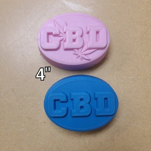 Puede incluir: Dos barras de jabón ovaladas, una rosa y otra azul, cada una con la palabra "CBD" en relieve en la parte superior. La barra de jabón rosa tiene un diseño de hoja alrededor de la palabra "CBD". Las barras de jabón tienen un diámetro de 10 cm.
