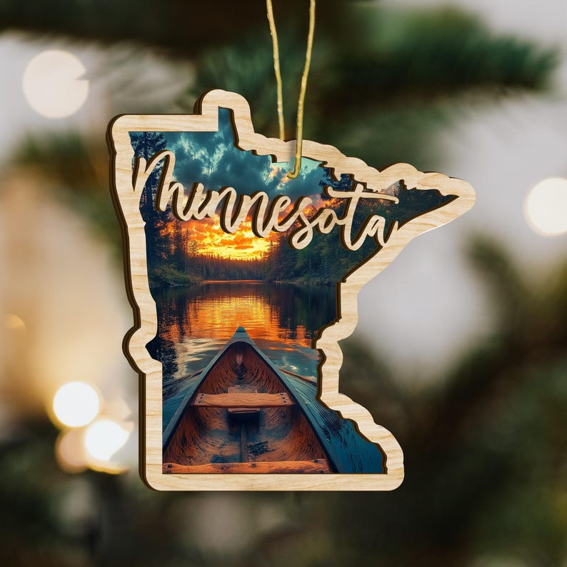 May include: Wooden ornament in the shape of the state of Minnesota, featuring a scenic lake view with a canoe and sunset. The word "Minnesota" is written across the top in a cursive font. A gold string is attached for hanging.