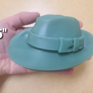 Könnte beinhalten: Ein grüner Fedora-Hut mit 12,7 cm Durchmesser, breiter Krempe und Schleife am Band.