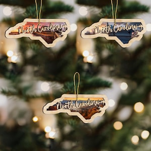 May include: Three wooden North Carolina ornaments with scenic landscape images inside. Each ornament is shaped like the state and has the words "North Carolina" cut out. The ornaments hang from gold strings.