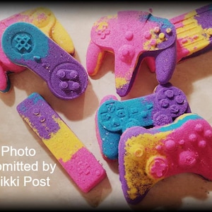 May include: A set of six colorful bath bombs shaped like video game controllers. The bath bombs are pink, yellow, blue, and purple.