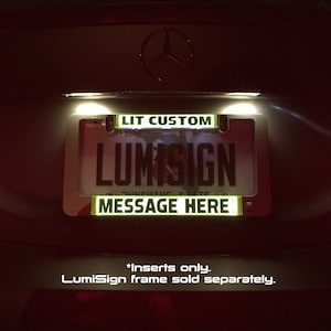 Op de afbeelding: Een close-up van een aangepaste kentekenplaathouder met de tekst "LIT CUSTOM" en "LUMISIGN" in witte letters op een zwarte achtergrond. De houder heeft ook de tekst "MESSAGE HERE!" in groene letters op een zwarte achtergrond. De houder is verlicht met witte LED-verlichting. *Alleen inzetstukken. LumiSign-frame apart verkrijgbaar.
