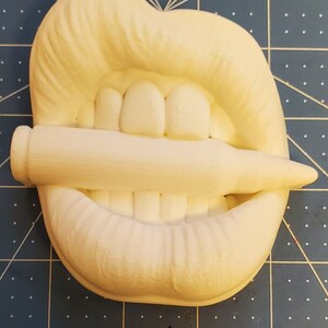 Pu&ograve; includere: Scultura bianca stampata in 3D di una bocca con i denti che mordono un proiettile. La scultura &egrave; su un tappetino da taglio blu.