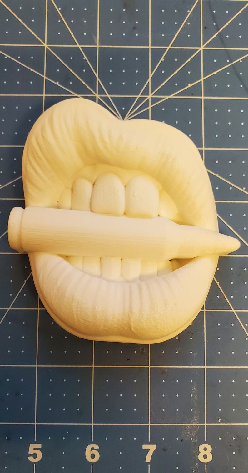 Pu&ograve; includere: Scultura bianca stampata in 3D di una bocca con i denti che mordono un proiettile. La scultura &egrave; su un tappetino da taglio blu.