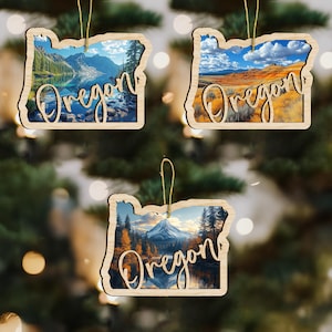 May include: Three wooden Oregon-shaped ornaments with scenic landscape images. Each ornament features a different landscape: a lake and mountains, rolling hills, and a mountain with a lake. The word "Oregon" is written in a cursive font on each ornament.