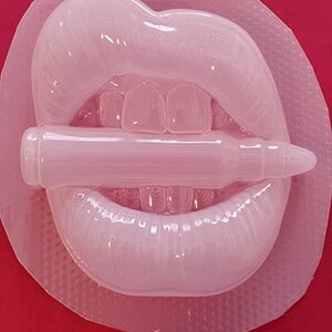 Pu&ograve; includere: Uno stampo in silicone bianco di un paio di labbra che mordono un proiettile. Lo stampo &egrave; su una superficie rossa.