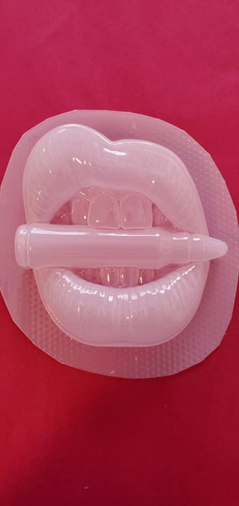 Pu&ograve; includere: Uno stampo in silicone bianco di un paio di labbra che mordono un proiettile. Lo stampo &egrave; su una superficie rossa.