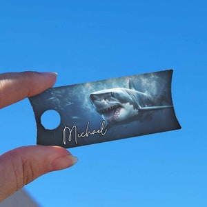 May include: A black and white personalised gift tag featuring a photograph of a great white shark swimming in the ocean. The tag has a hole punched at the top and the name "Michael" written in white cursive.