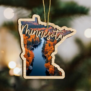 May include: Wooden ornament in the shape of Minnesota, featuring a scenic landscape of a river surrounded by vibrant orange autumn trees. The word "Minnesota" is scripted across the image. A gold string is attached for hanging.