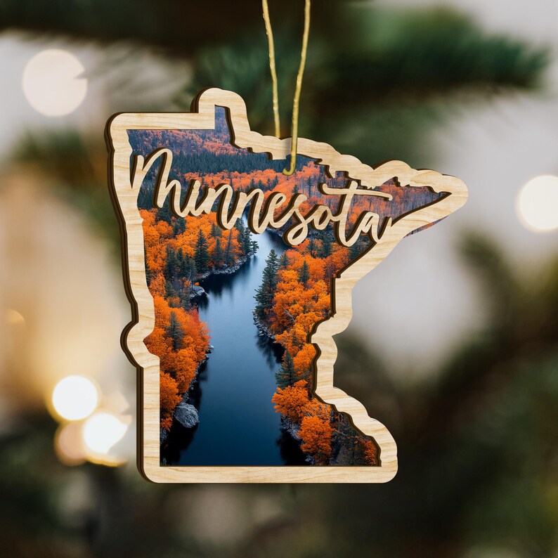 May include: Wooden ornament in the shape of Minnesota, featuring a scenic landscape of a river surrounded by vibrant orange autumn trees. The word "Minnesota" is scripted across the image. A gold string is attached for hanging.