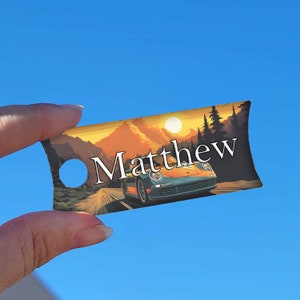 May include: A brown, orange, and blue car drives through a sunset landscape with a mountain in the background. The car is a Porsche 911. The name "Matthew" is printed on the tag.