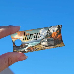 May include: A colourful illustrated tag with the name "Jorge" printed on it. The tag features a landscape scene with a white camper van driving through a forest.