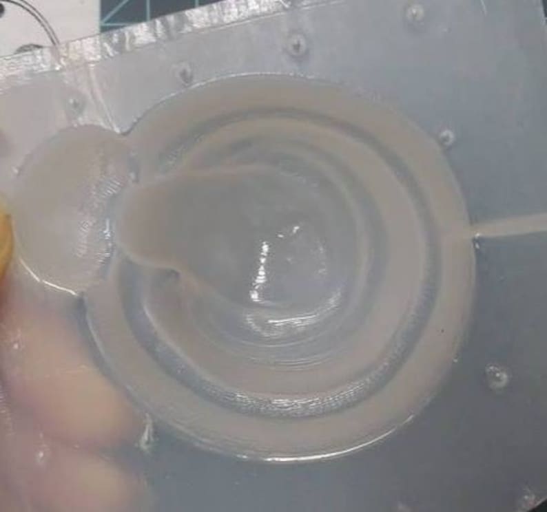 Op de afbeelding: Een close-up van een wit, rond, 3D-geprint object met een verhoogd, concentrisch ringontwerp. Het object is gedeeltelijk gevuld met een witte, viskeuze substantie.